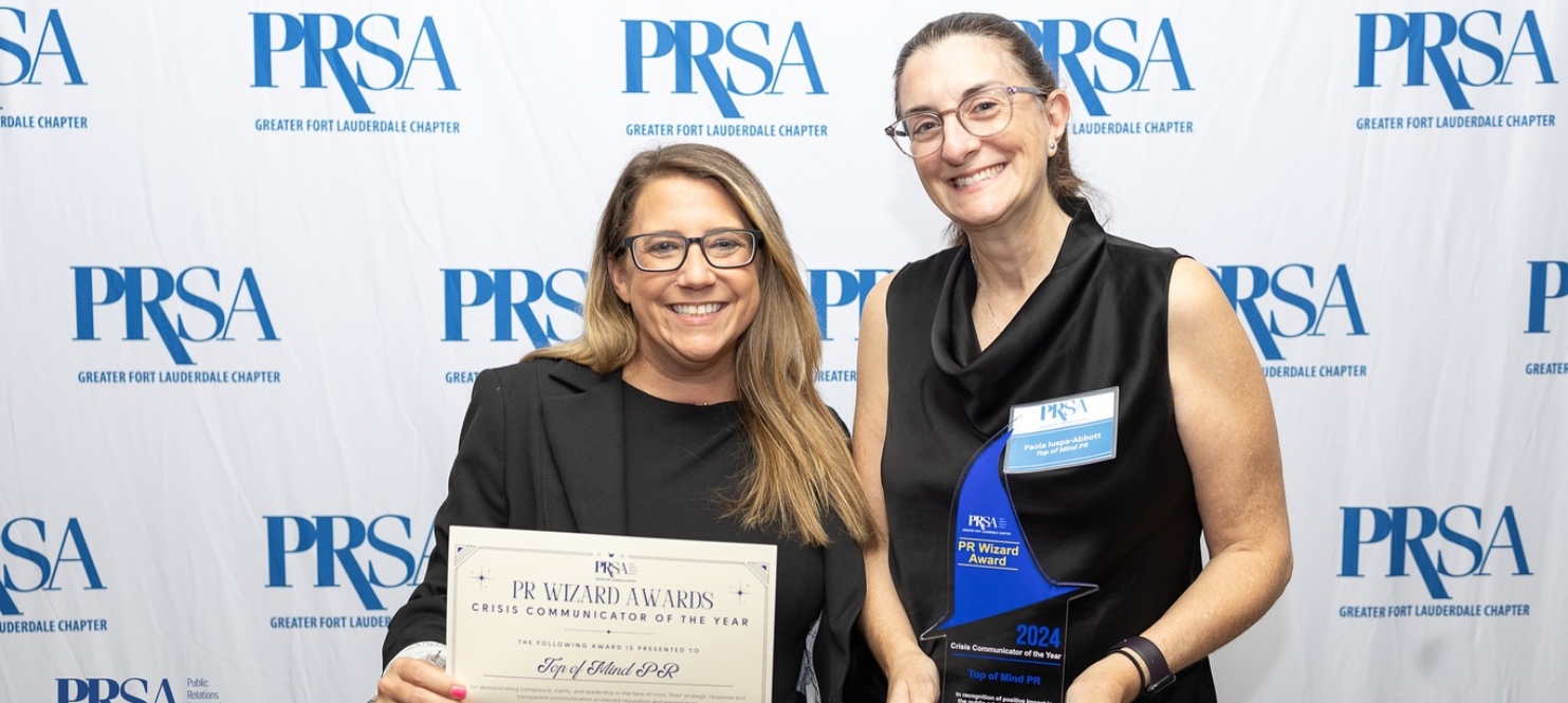 Paola Iuspa-Abbott and Jessica Forres standing together at a PRSA (Public Relations Society of America) event. One woman is holding a certificate labeled "PR Award Winner" while the other is holding a trophy. Both are smiling, and they are standing in front of a backdrop displaying the PRSA logo