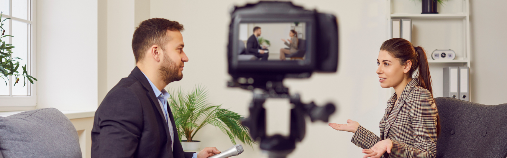 A man and woman sit across from each other during a recorded interview in a modern office setting