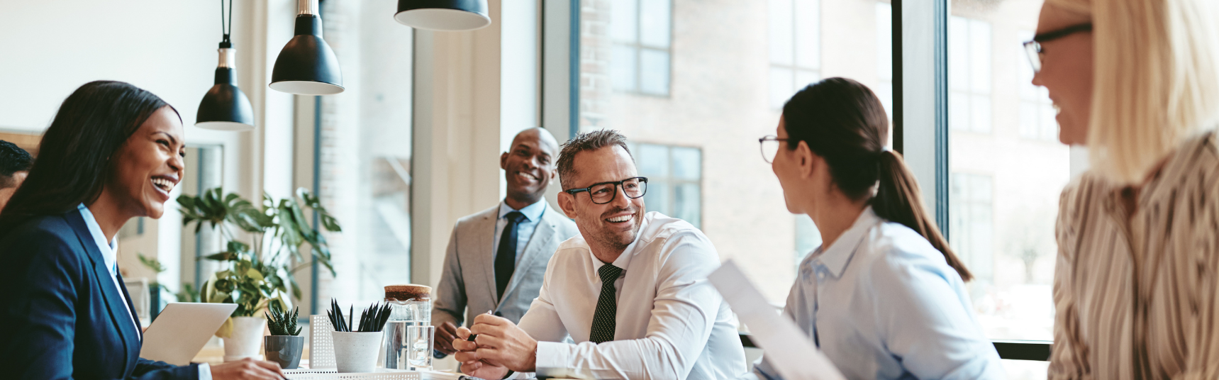 A group of professionals sitting around a conference table in a modern office smiling and engaging in a collaborative meeting Natural light fills the room through large windows creating a bright and welcoming atmosphere