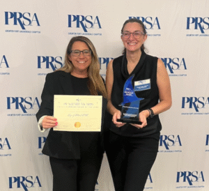 Paola Iuspa Abbott and Jessica Forres standing together at a PRSA (Public Relations Society of America) event. One woman is holding a certificate labeled "PR Award Winner" while the other is holding a trophy. Both are smiling, and they are standing in front of a backdrop displaying the PRSA logo