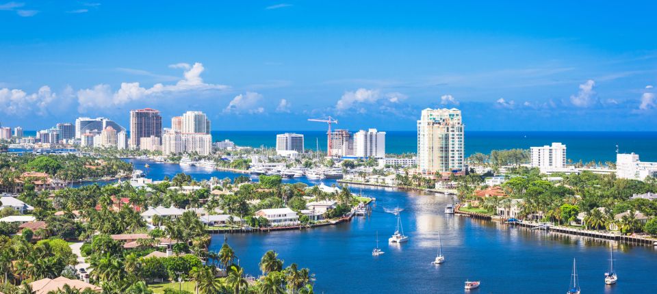 Scenic view of Fort Lauderdale with tall buildings along the coastline a waterway with boats and yachts surrounded by palm trees and residential houses under a bright blue sky