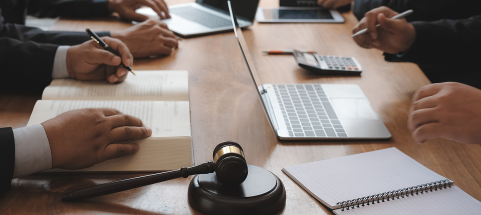 Close up of people in business attire sitting around a wooden table with open notebooks laptop calculator and a gavel suggesting a legal meeting or law firm discussion