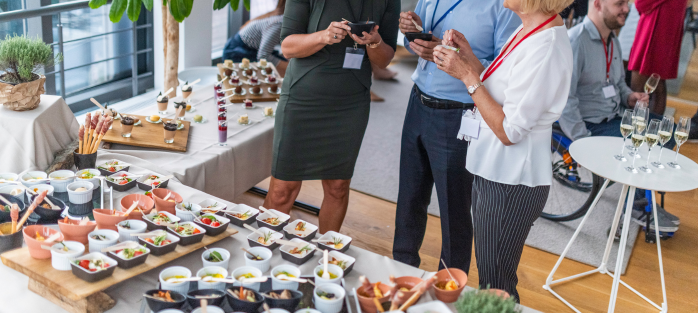 Group of people socializing at an indoor brand activation while standing near a large table covered with assorted appetizers small plates and glasses of champagne