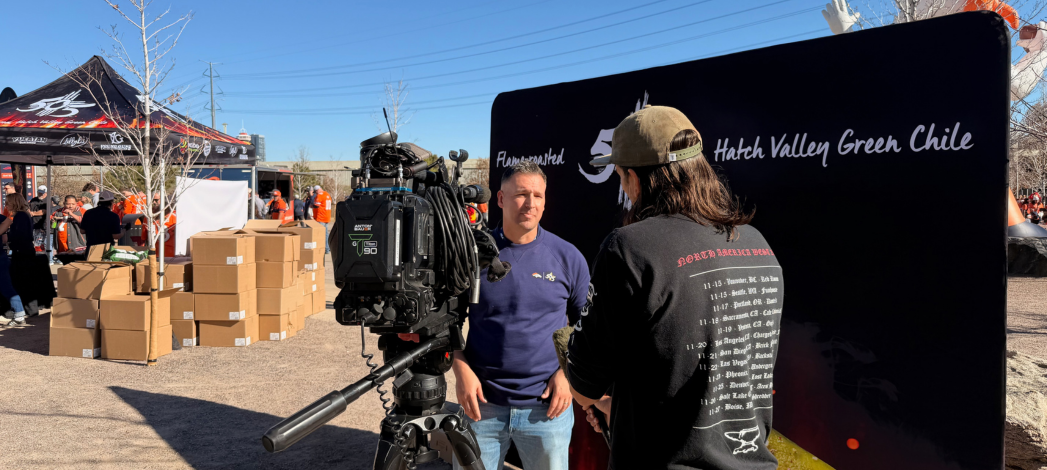Outdoor brand activation event where a reporter interviews a man on camera surrounded by stacked boxes branded tents and event staff under a clear blue sky
