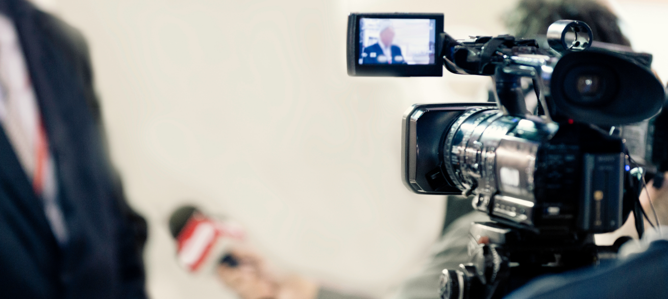 Video camera recording an interview with a spokesperson during a media interview. The image represents television news, media relations, public relations, press coverage, broadcast journalism, and corporate communications.