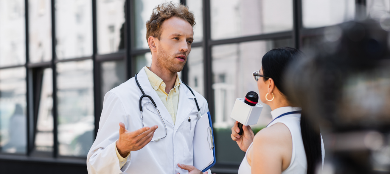 A television reporter interviews a doctor holding a clipboard outside a medical facility.