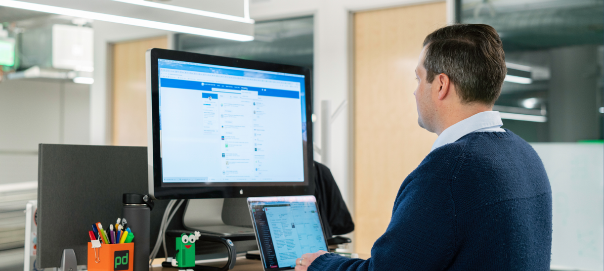 Employee working at a standing desk in a modern office, using a laptop and a large external monitor displaying a professional networking website. Office supplies, a reusable water bottle, and desk accessories are visible in the workspace, illustrating a business professional engaged in online networking or business development.